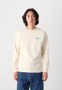 Crème-kleurige sweatshirt met lange mouwen, ribgebreide boorden en zoom. Heeft een klein lichtblauw vierkant iets op de borst. Soepele textuur.
