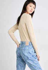 Suéter de malha bege com mangas longas, combinado com jeans de denim azul de cintura alta e desgastados, apresentando um logo de couro na parte de trás.