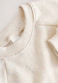 Sweat-shirt crème clair avec col et poignets côtelés, présentant de petites taches multicolores réparties sur tout le tissu. Fini doux et texturé.