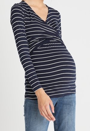 Personne enceinte portant un haut à manches longues rayé bleu marine et blanc et un jean bleu sur un fond uni.