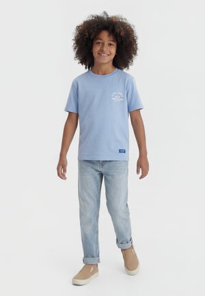 Boy with curly hair stands smiling, wearing a light blue graphic T-shirt, light-wash jeans rolled at the cuffs, and beige slip-on shoes.