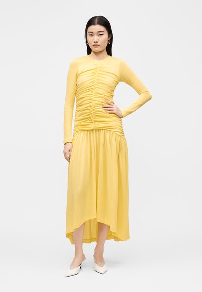 Femme debout, main sur la hanche, portant une robe jaune froncée à manches longues et des chaussures blanches pointues sur un fond clair uni.