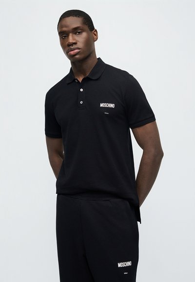 Νέος άνδρας που φοράει μαύρο πόλο Moschino και αντίστοιχο παντελόνι, στέκεται μπροστά σε απλό ανοιχτό γκρι φόντο.