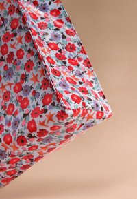 Közelkép egy szövet táska sarkáról, mely élénk virágmintával díszített, piros, lila és narancssárga virágokkal világos alapon.