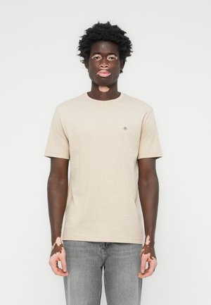 Beige bomull t-skjorte med korte ermer, rund hals, og en liten brodert logo på venstre bryst. Modellen har på seg falmede grå jeans.