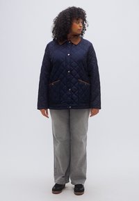 Μπλε ναυτικό μπουφάν με καπιτονέ σχέδιο, καφέ γιακά και τσέπες, με κουμπιά τύπου snap και μακριά μανίκια. Συνδυασμένο με γκρι τζιν και μαύρα αθλητικά παπούτσια.