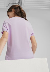 Liliowy T-shirt z krótkim rękawem o gładkiej fakturze. Prezentuje luźny krój i posiada okrągły dekolt. Włosy w warkoczu.
