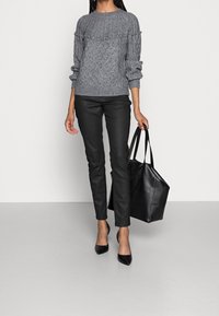 Žena oblečená v šedém pleteném svetru s texturou, černých přiléhavých kalhotách, černých lodičkách s špičatou špičkou, držící velkou černou koženou tašku přes rameno.