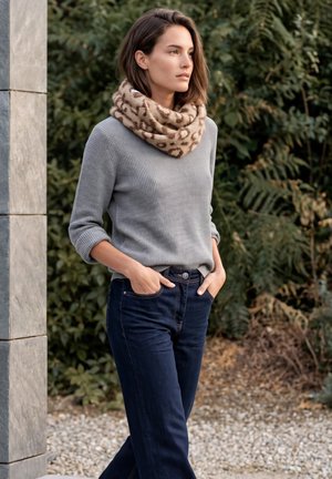 Une femme portant un pull gris, une écharpe à imprimé léopard et un jean foncé se tient dehors, les mains dans les poches.