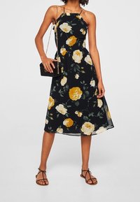Robe midi florale en tissu noir avec des roses jaunes et blanches. Dotée de bretelles spaghetti, d'un corsage ajusté et d'une jupe fluide et ample.