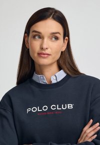 Mujer con cabello largo castaño, usando pequeños pendientes de aro, una camisa a cuadros azul y una sudadera oscura con el texto "POLO CLUB ESTABLISHED 2012".