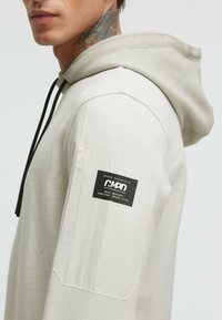 Light gray hoodie with textured fabric, a drawstring hood, and a black label stitched on the left sleeve. Side view displays shoulder seam.