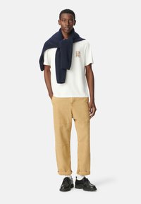 Camiseta blanca con "LDN PRS" en marrón, pantalones de pierna ancha beige, suéter azul marino sobre los hombros y zapatos negros con suelas gruesas.