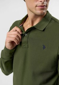 Zelená polo košeľa z textúrovanej tkaniny, s dvoma gombíkmi na lége a malým námorníckym logom na hrudi. Červené a biele akcenty vo vnútri goliera.