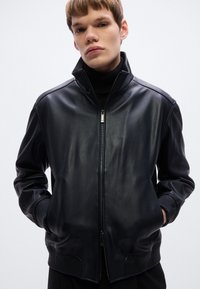 Fekete bőr bomberdzseki cipzáras elülső résszel, bordázott mandzsettákkal és szegéllyel, valamint magas gallérral, sima, elegáns textúrával.