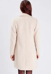 Béžový kabát z vlněné směsi s rovným střihem, rozparkem vzadu a dlouhými rukávy. Má klopový límec a boční kapsy.