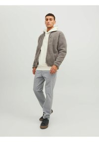 Giacca in pile grigia con chiusura a zip, indossata sopra un hoodie bianco. Pantaloni grigi a righe e sneakers nere completano l'outfit.
