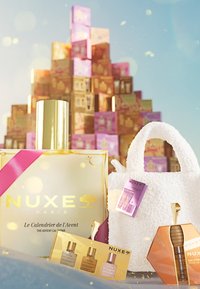 Botella dorada con una cinta rosa, bolsa blanca texturizada y varias cajas de productos NUXE en colores pastel en el fondo.
