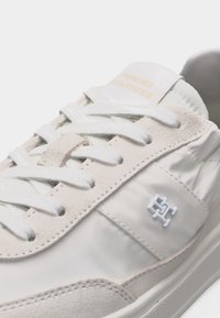 Baskets blanches et beiges avec des lacets blancs, des panneaux en cuir suédé et en tissu lisse, et un petit logo blanc en relief sur le côté.