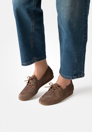 Feet wearing brown lace-up moccasin shoes and cropped blue jeans on a white background.