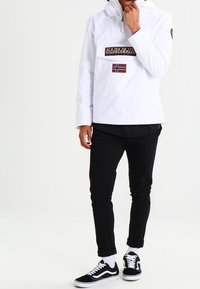 Chaqueta blanca tipo pulóver con cremallera parcial y parche con logo, combinada con pantalones ajustados negros y zapatillas negras y blancas.