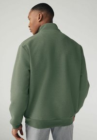 Veste verte texturée avec un col montant et des poignets côtelés. Le tissu présente un motif en losanges et une couleur unie au niveau de l'ourlet.