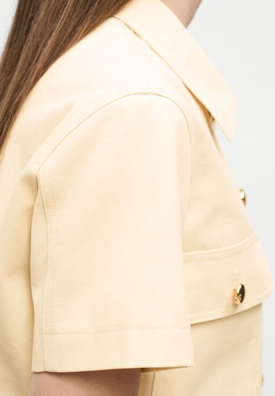 Κοντινό πλάνο ενός ατόμου που φοράει μπεζ κοντομάνικο σακάκι με χρυσά κουμπιά και μυτερό γιακά, με ορατά ανοιχτό καστανά μαλλιά.