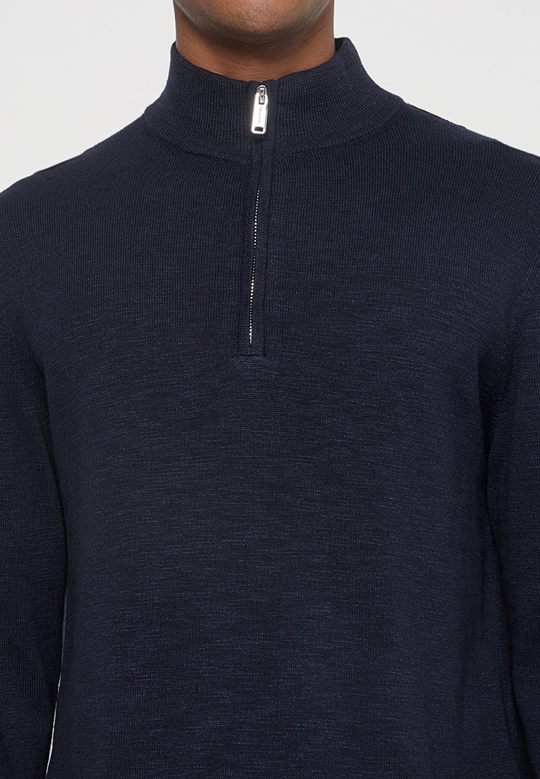 Mężczyzna w ciemnogranatowym swetrze dzianinowym z kołnierzem na ćwierć-zamek, widoczny od ramion do podbródka.