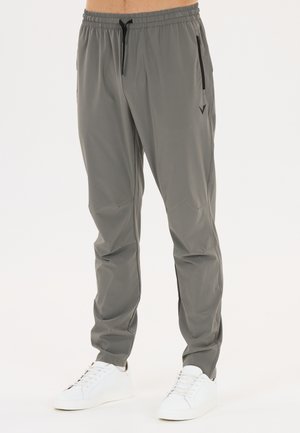 Homme portant un pantalon gris à cordon avec poches latérales zippées et des baskets blanches, debout devant un fond clair uni.