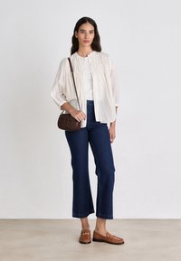 Blusa beige chiaro con bottoni e texture a coste, abbinata a jeans a campana blu scuro e mocassini marroni. È inclusa una piccola borsa a tracolla marrone trapuntata.