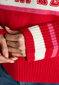 Red sweater with ribbed texture, featuring horizontal stripes in darker and lighter red, pink, and white. Cuffs are also ribbed.