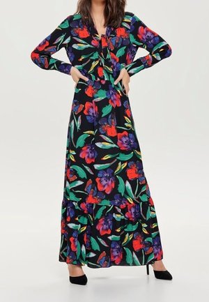 Žena v dlouhých černých šatech s velkými květinovými vzory v červené, fialové a zelené barvě, doplněná černými botami na vysokém podpatku.