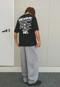 Černé oversized tričko s bílým grafickým textem na zádech. Spárované se světle šedými volnými tepláky a černou obuví.