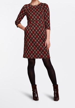 Femme portant une robe à carreaux rouge mi-longue avec des manches trois-quarts, des collants noirs et des bottines marron foncé brillantes, debout avec une main dans la poche.