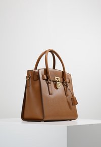Borsa in pelle marrone con doppio manico, lucchetto dorato frontale e forma rettangolare strutturata su una superficie bianca contro uno sfondo semplice.