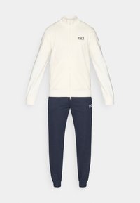 Krēmkrāsas džemperis ar rāvējslēdzēju un augstu apkakli, kombinēts ar tumši zilām sporta biksēm. Džempera kreisajā krūtīs ir logotips; biksēm kreisajā augšstilbā arī ir logotips.