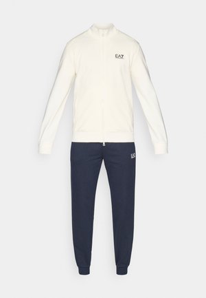 Krem zip-up jakke med høy krage, kombinert med marineblå joggebukser. Jakken har logo på venstre bryst; buksene har logo på venstre lår.