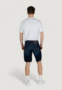 Shorts en denim bleu foncé avec des coutures jaunes, dotés d'une taille et de deux poches arrière. Associés à un t-shirt blanc uni et des baskets blanches.