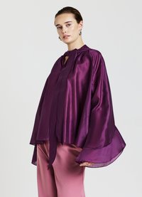 Blusa di raso viola con vestibilità ampia, colletto con fiocco e maniche svasate, abbinata a pantaloni rosa chiaro. Texture liscia e design fluido.