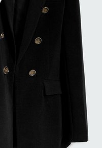 Manteau noir croisé à double boutonnage avec six boutons décoratifs dorés et une poche à rabat sur le côté droit.