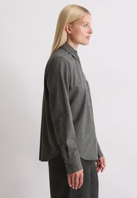 Γκρι πουκάμισο με κουμπιά, φτιαγμένο από υφασμάτινη υφή, με γιακά, μακριά μανίκια και χαλαρή εφαρμογή. Είναι ορατή και μια τσέπη στο στήθος.