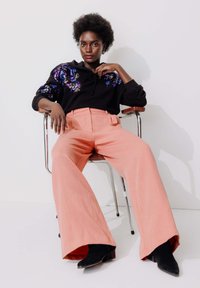 Sweat à capuche noir avec un design floral en sequins, associé à un pantalon large rose clair et des bottines noires, assis sur une chaise en métal.