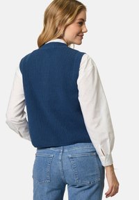 Dunkelblauer Strickwesten mit geripptem Muster, tailliertem Schnitt und breiten Schulteröffnungen, getragen über einem weißen Hemd mit gepunkteten Akzenten.