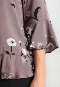 Blommig blus i mauve tyg med klockärmar, med vita och svarta blommönster och en volangdetalj vid nederkanten.