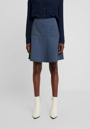 Woman wearing a dark blue blouse, textured blue A-line skirt, and white ankle boots, standing against a plain light background.