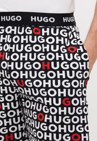 Shorts noirs avec un motif répétitif du mot "HUGO" en lettres blanches et rouges. Taille élastique avec le logo "HUGO". Matériau en coton.