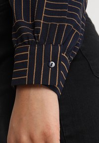 Polsino di camicia blu navy con strisce orizzontali dorate, dotato di un unico bottone blu navy e di un tessuto testurizzato. Primo piano del polso su uno sfondo scuro.