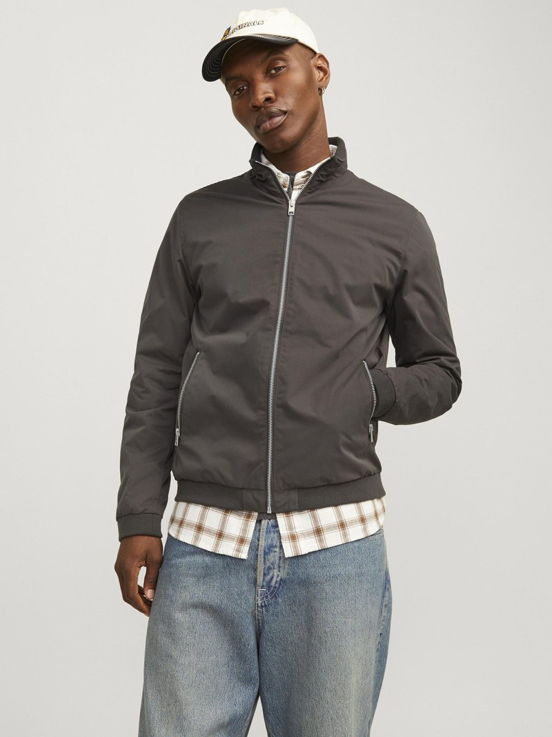 Black zip-up jacket with a high collar, featuring side pockets and ribbed cuffs, worn over a checkered shirt and paired with light blue jeans.
