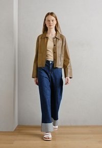 Veste en cuir marron cropped, top beige, jeans en denim noir à jambes larges avec des poignets bleu clair, et sandales blanches. Modèle marchant sur un fond neutre.