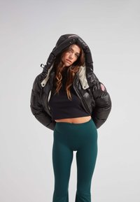 Chaqueta de plumas negra con un forro blanco y capucha oversize, con cierre de cremallera y un detalle en color rosa. Combinada con leggins verdes.